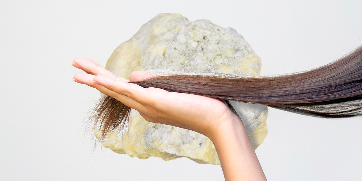 mano vista laterale che mantiene ciocca di capelli con foto di un minerale di zolfo sullo sfondo