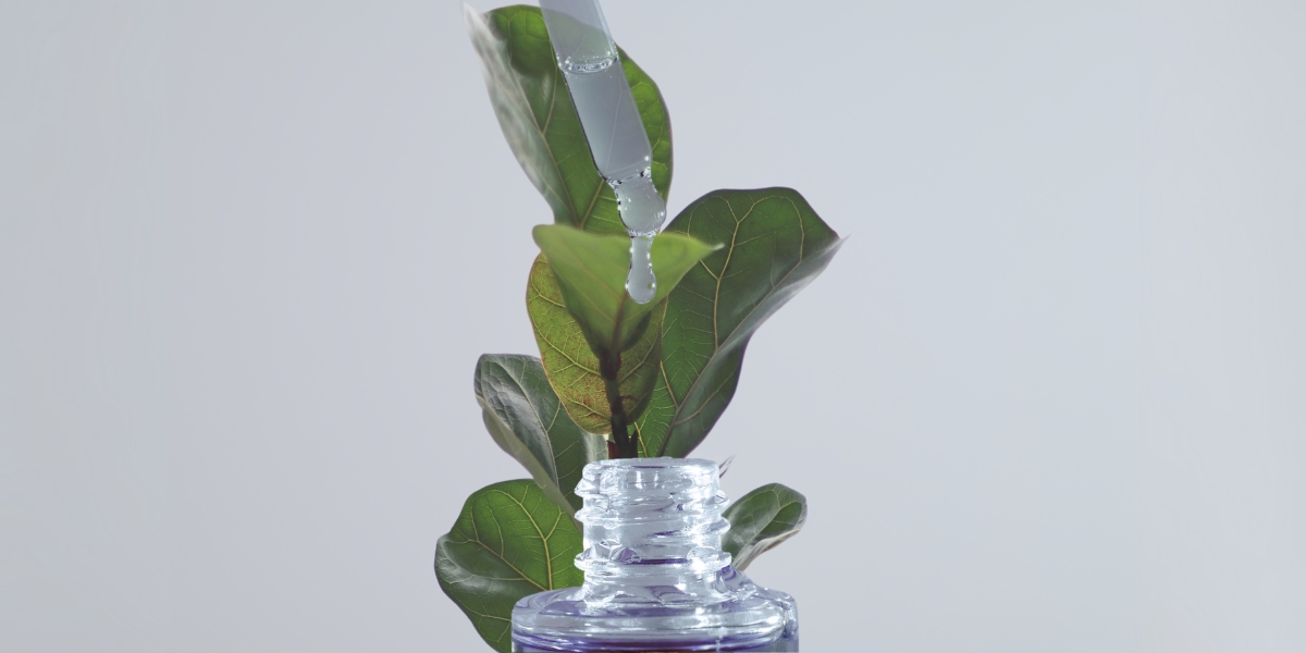 foto di un contagocce in alto dal quale cola una goccia verso il basso dov'è posizionato il collo di una bottiglietta con una pianta di fico dietro su uno sfondo grigio